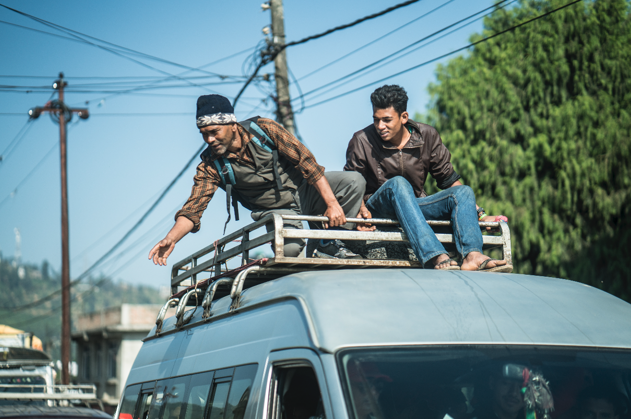 Two men riding on top of a small minivan in Nepal, one is reaching down to someone.